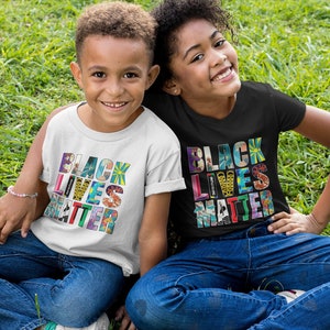 May include: Two children wearing white and black t-shirts with the phrase "Black Lives Matter" printed in colorful letters. The children are sitting on a grassy surface.