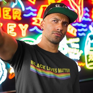 May include: A person wearing a black t-shirt with the text "Black Lives Matter" in yellow letters and a rainbow stripe below the text. The person is wearing a green baseball cap with the text "ST 20" on it.