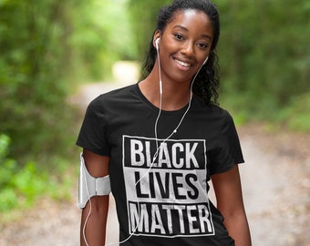 May include: A black t-shirt with a white graphic that says "Black Lives Matter" in a distressed font. The shirt is being worn by a person with dark skin and curly hair. The person is standing on a path in a forest.
