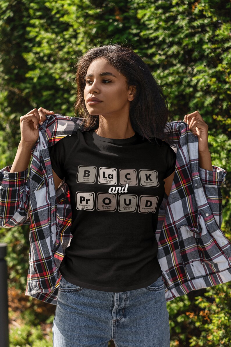 May include: Black t-shirt with the words "Black and Proud" spelled out using periodic table elements. The shirt is black with white text. The person in the image is wearing a plaid shirt over the t-shirt and blue jeans.