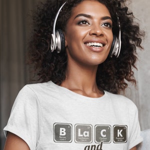 May include: A light gray t-shirt with the words "Black and Proud" spelled out using elements from the periodic table. The person in the image is wearing silver headphones.
