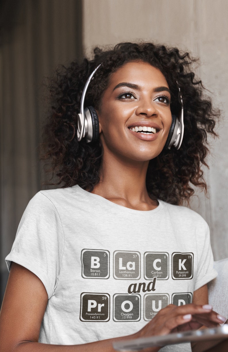 May include: A light gray t-shirt with the words "Black and Proud" spelled out using elements from the periodic table. The person in the image is wearing silver headphones.