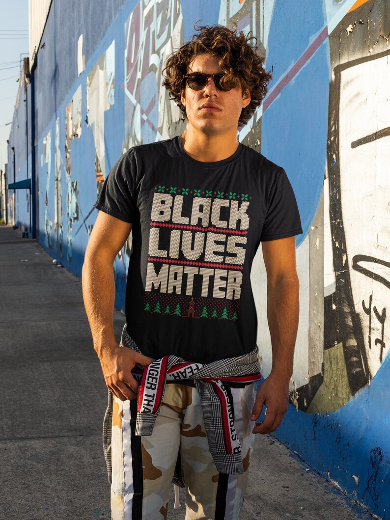 May include: A person wearing a black t-shirt with the text "Black Lives Matter" in white letters with a red and green Christmas-style pattern. The person is wearing camouflage pants with a black stripe down the side and a black and white plaid shirt tied around their waist.