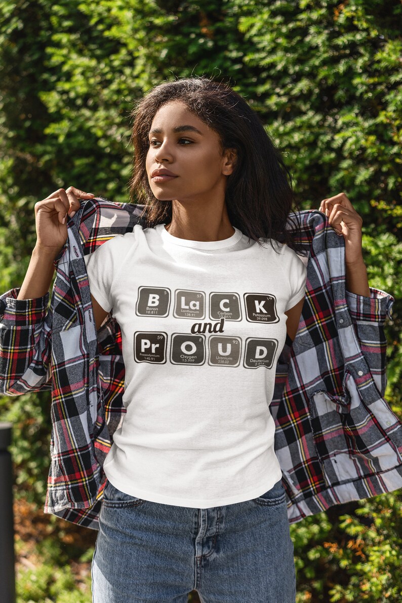 May include: White t-shirt with the text "BLACK and PROUD" in a periodic table style. The shirt is worn with a plaid shirt and blue jeans. The background is a green, leafy area.