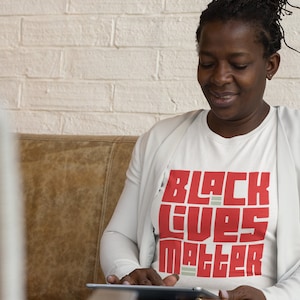 May include: A woman wearing a white t-shirt with the words "Black Lives Matter" printed in red block letters. She is sitting on a brown leather couch and looking down at a tablet.