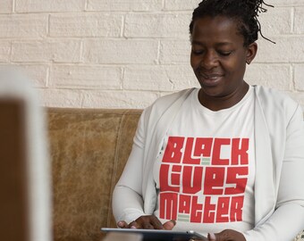 May include: A woman wearing a white t-shirt with the words "Black Lives Matter" printed in red block letters. She is sitting on a brown leather couch and looking down at a tablet.