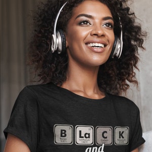 May include: A black t-shirt with the words "Black and Proud" spelled out using periodic table elements. The shirt is worn by a person wearing silver headphones.