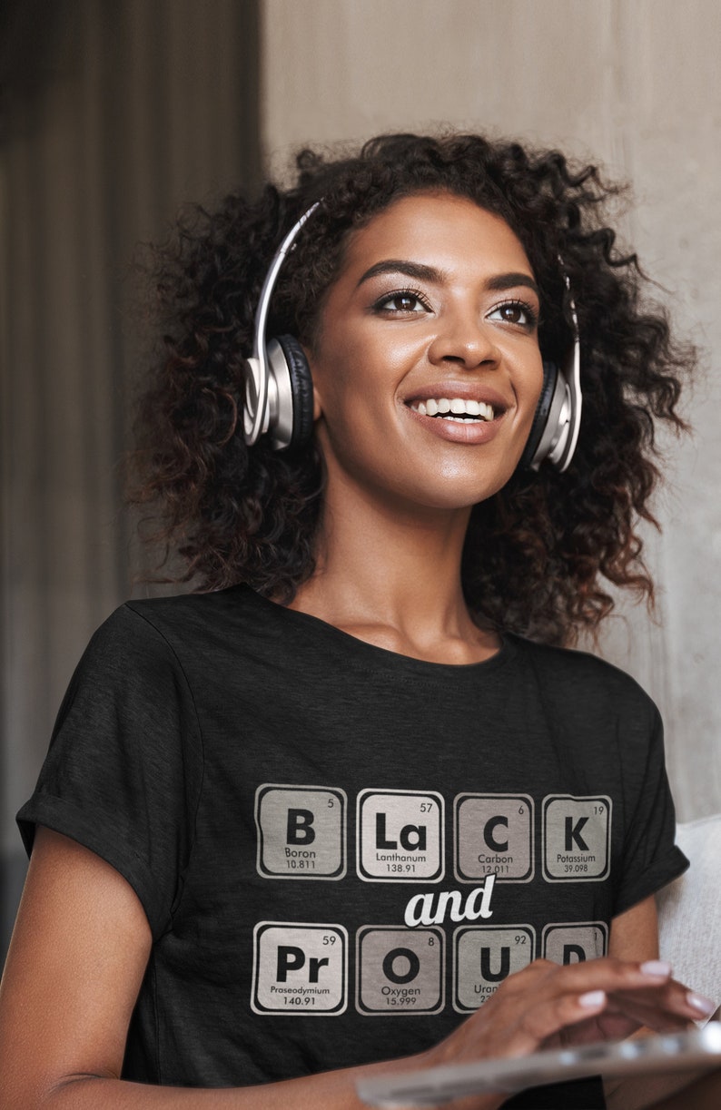 May include: A black t-shirt with the words "Black and Proud" spelled out using periodic table elements. The shirt is worn by a person wearing silver headphones.