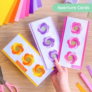 May include: Three aperture cards with floral designs in yellow, purple, and pink, each with a gold glitter center. The cards are on a wooden surface with colorful paper strips and scissors.
