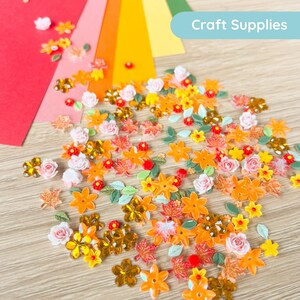 May include: A selection of craft supplies, featuring orange, yellow, and pink flower and leaf-shaped embellishments. The image also shows colourful paper in red, pink, yellow, and green. The text "Craft Supplies" is visible in a light blue banner.