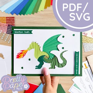 May include: A green papercraft dragon with a red and yellow flame coming out of its mouth. The dragon is on a white card with a green border. The card has the text "Adventure Awaits" on it. The card is being held by a hand. The image also includes a sheet of paper with the text "Dragon Trainer" on it.