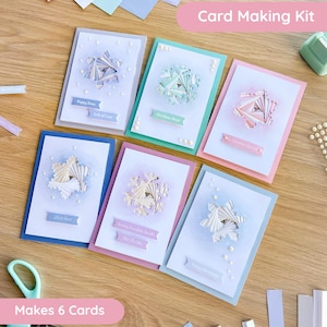 May include: A card making kit featuring six handmade greeting cards with intricate paper snowflake designs. Each card displays unique pastel colors, small white embellishments, and printed banners with text including Happy Xmas, Christmas Magic, and Let It Snow.