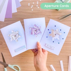 May include: Three white greeting cards featuring intricate paper-folded snowflake apertures in shades of purple, blue, and white. The cards display snowflake patterns, small white dots, and text banners reading Season's Greetings and Let it Snow.