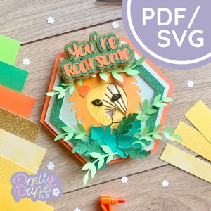 Puede incluir: Un diseño de león de papel en capas con el texto "You're Roarsome". El león es naranja y amarillo, rodeado de hojas verdes y un marco hexagonal verde y naranja. La imagen incluye recortes de papel y el texto "PDF/SVG".