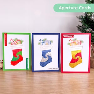 Puede incluir: Tres tarjetas de Navidad con diseño de calcetín. Cada tarjeta tiene un borde de diferente color: verde, azul y rojo. Los calcetines están llenos de regalos y bastones de caramelo. El texto "Aperture Cards" está en la parte superior.
