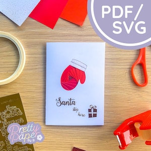 May include: A white card with a red paper-quilled mitten, a gift box, and the text "Santa stop here". The card is on a wooden surface with craft supplies.