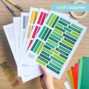 May include: A selection of Christmas-themed sticker sheets featuring festive messages in green, red, and white. The stickers are on white paper, held by a hand. The words "Craft Supplies" are at the top. Various colored paper and craft supplies are also visible.