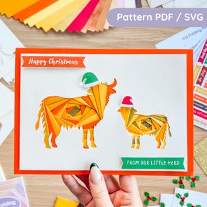 May include: A handmade Christmas card featuring two origami-style cows wearing Santa hats. The card has the text "Happy Christmas" and "From Our Little Herd" on it. The card is orange and white with a red border.