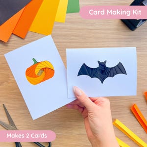 May include: A card making kit with two white cards, one featuring a pumpkin design and the other a bat. The pumpkin is orange and yellow, and the bat is black. The text "Card Making Kit" and "Makes 2 Cards" are visible.