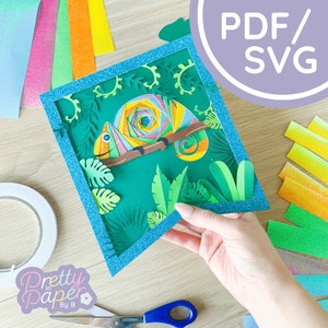 May include: A blue frame with a green background featuring a colorful paper chameleon with a swirl pattern. The chameleon is on a branch with green paper leaves around it. The text "PDF/SVG" is in a purple circle on a white background.