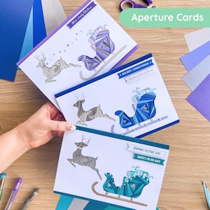 Puede incluir: Tres tarjetas navideñas hechas a mano con un diseño de reno y trineo. Las tarjetas presentan un fondo blanco con detalles en azul, morado y turquesa. El texto en las tarjetas dice "Have a lovely Christmas", "Merry Christmas" y "Sending Festive Joy". Las tarjetas están etiquetadas como "Aperture Cards".