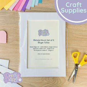 May include: A white picture mount set of three, packaged in clear plastic. The text "Picture Mount Set of 3 Bright White" is visible. The outer edge is A4 size, approximately 8.27 inches wide and 11.69 inches high. A pair of yellow scissors is on the right.