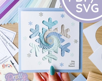 May include: A white card with a blue and white snowflake design. The snowflake is made of paper and has a 3D effect. The card also has the words "Joy Love Peace" written on it.
