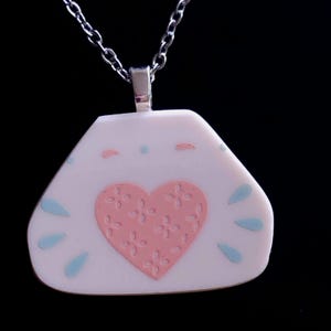 Puede incluir: Collar con colgante blanco con un corazón rosa con diseño floral. Gotas azules y puntos rosas decoran el fondo blanco. La cadena es plateada.