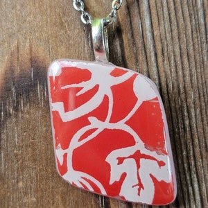 May include: A silver chain necklace with a red and white pendant. The pendant is a geometric shape with a red background and a white floral design. The necklace is displayed on a wooden surface.