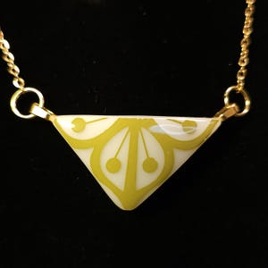 Puede incluir: Un collar de cadena dorada con un colgante triangular. El colgante es blanco con un diseño floral amarillo-verdoso. El collar se muestra sobre un fondo negro.