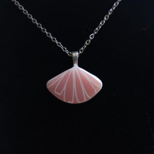 Op de afbeelding: Een zilveren ketting met een roze hanger. De hanger is waaiervormig met witte lijnen die vanuit het midden uitstralen. De achtergrond is zwart.