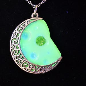 May include: A silver crescent moon pendant with a green glow-in-the-dark resin moonstone and a green rhinestone in the centre.