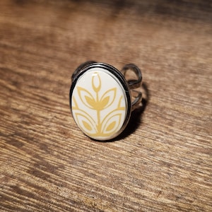 May include: Silver-toned adjustable ring with an oval, cream-colored centerpiece. The centerpiece features a yellow floral design. The ring is set against a wooden background, showcasing the jewelry's details.