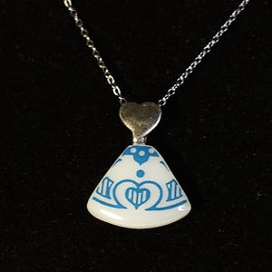 Puede incluir: Un collar de plata con un colgante con base blanca y diseño decorativo azul. El colgante tiene una parte superior en forma de corazón y una forma triangular con un corazón azul y un diseño de corona. El collar está sobre un fondo negro.