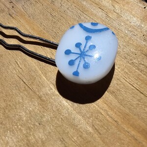 Pode incluir: Um grampo de cabelo branco com um design de floco de neve azul. O grampo tem formato oval e um acabamento brilhante.