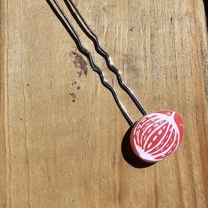 May include: A pair of silver hair pins with a red and white patterned ceramic end cap. The pattern resembles a flower or a seed pod.