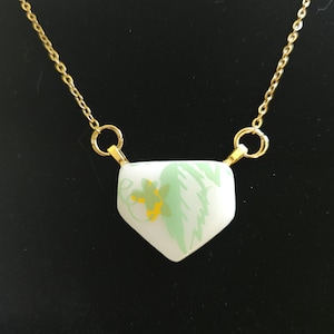 May include: A gold-colored chain necklace with a white pendant. The pendant is shield-shaped and decorated with green and yellow floral designs. The necklace is displayed against a black background.