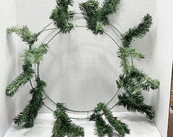 May include: A green wire wreath form with green pine sprigs attached to it. The wreath is laying flat on a white surface.
