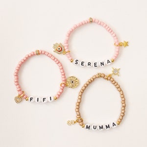 May include: Three beaded bracelets with charms and name spellings. The bracelets are pink and gold with white letter beads spelling "SERENA", "FIFI", and "MUMMA".