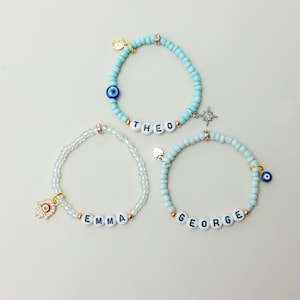 Puede incluir: Tres pulseras de cuentas con los nombres "THEO", "EMMA" y "GEORGE". Las pulseras presentan varios dijes, incluyendo un ojo turco, una estrella, un corazón y una mano de Fátima. Las cuentas son en tonos azules, blancos y transparentes.