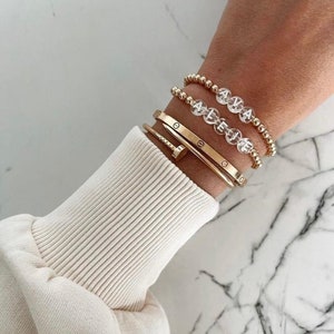 Può includere: Un braccialetto d'oro con il nome "Ava" scritto in perle trasparenti. Il braccialetto è indossato su un polso con un braccialetto rigido d'oro e un braccialetto a catena d'oro.