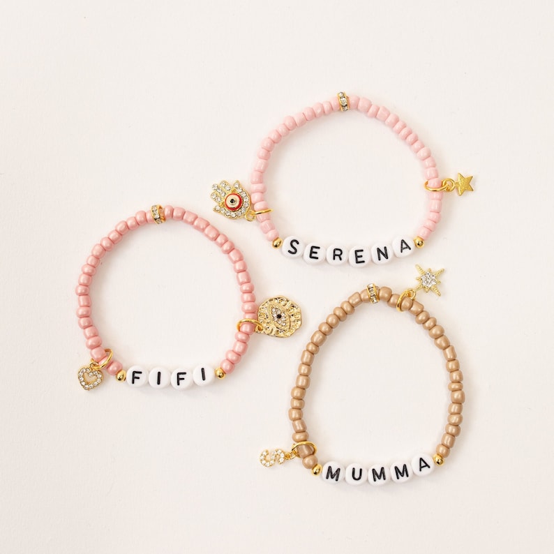 May include: Three beaded bracelets with charms and personalized names. The bracelets are pink and gold with charms including a hamsa hand, an evil eye, a star, and a heart. The names on the bracelets are "Fifi", "Serena", and "Mumma".
