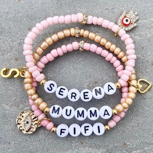 May include: Three layered bracelets with pink and gold beads. The bracelets have charms including a heart, an evil eye, and letter beads spelling out the names "SERENA", "MUMMA", and "FIFI".