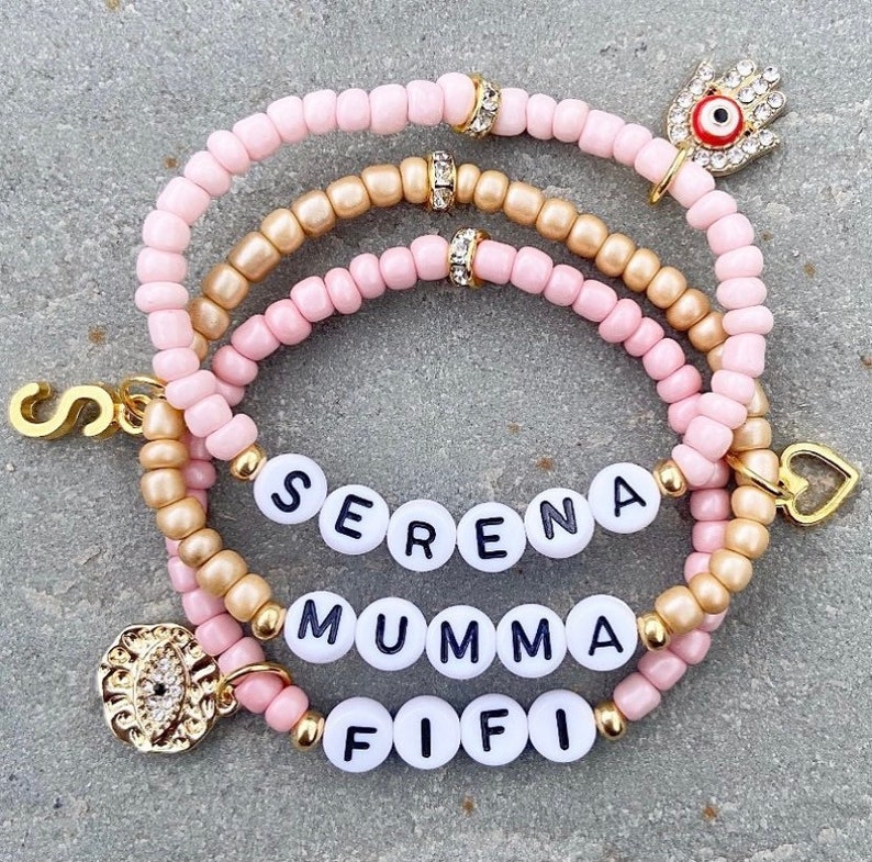 May include: Three layered bracelets with pink and gold beads. The bracelets have charms including a heart, an evil eye, and letter beads spelling out the names "SERENA", "MUMMA", and "FIFI".