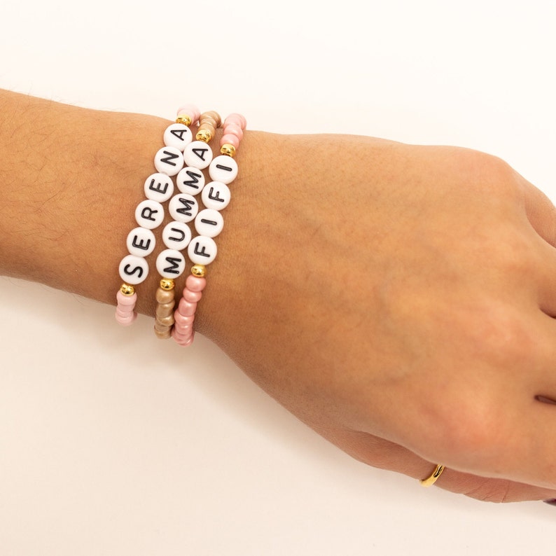 May include: Three beaded bracelets with the names "SERENA", "MUMMA", and "FIFI" spelled out in black letters on white beads. The bracelets are made with pink, gold, and white beads.