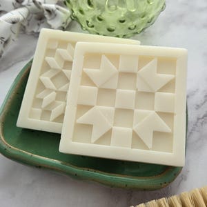 May include: Two square, off-white soaps with geometric patterns rest on a small, green ceramic dish. A green glass vase and a wooden brush are also visible. The soaps have a quilt-like design, perfect for a spa or bath-themed gift.