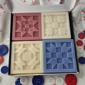 May include: A box set of four square soaps, each featuring a different quilt block design in shades of pink, cream, and blue. The soaps are surrounded by red, white, and blue buttons and a fabric banner with patriotic stars.