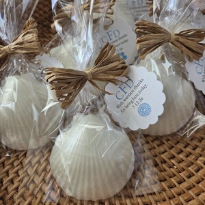 May include: Several seashell-shaped soaps wrapped in clear cellophane, tied with raffia bows, and tagged with the text "CFD With warmest thanks for being here today 7-12-26". The soaps are white and sit in a woven basket.