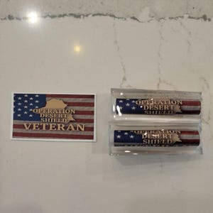 May include: An American flag design with the words "OPERATION DESERT SHIELD VETERAN" is displayed on a rectangular sticker. Two cylindrical items with the same design are encased in clear plastic.