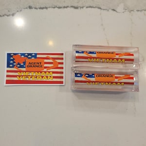 Puede incluir: Una pegatina de Veterano de Vietnam del Agente Naranja y dos pisapapeles de acrílico transparente. La pegatina presenta un diseño de bandera con un mapa naranja de Vietnam y el texto "AGENT ORANGE VIETNAM VETERAN". Los pisapapeles tienen un diseño similar.